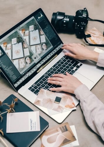Fotografė dirba su nuotraukų dizainu kompiuteryje, šalia guli fotoaparatas – kvietimas susipažinti su fotografijos kursais